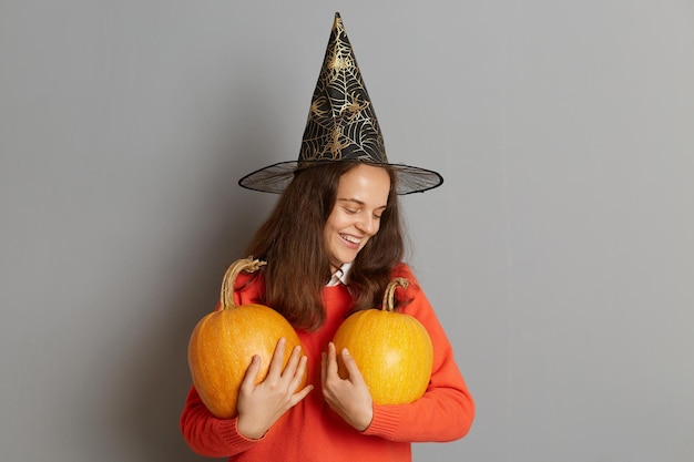 Zdjęcie zadowolona młoda dorosła kobieta w kapeluszu wiedźmy stojąca z dwoma pomarańczowymi dyniami w rękach na białym tle nad szarym tłem z okazji Halloween w dobrym świątecznym nastroju