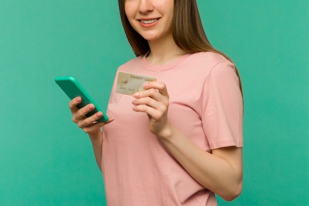 Zdjęcie zadowolona szczęśliwa krzycząca młoda kobieta pozowanie na białym tle na niebieskim tle ściany za pomocą telefonu komórkowego, trzymając kartę kredytową.