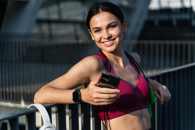 Zdrowa brunetka kobieta trzyma smartfon i szeroko uśmiecha się do kamery po ciężkim treningu na stadionie Koncepcja sportu