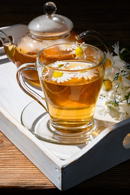 Zdrowa herbata ziołowa w filiżance i czajniczku, kwiaty rumianku na białej drewnianej tacy