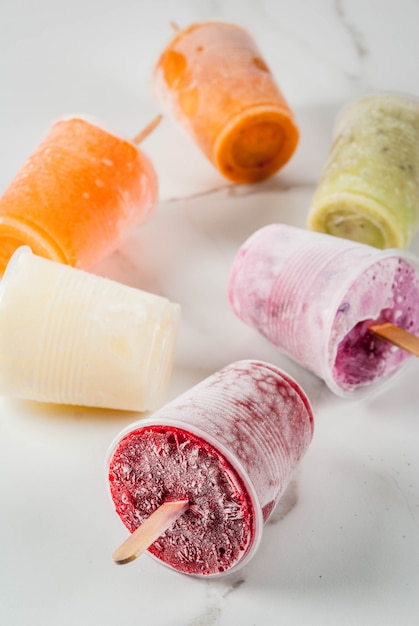 Zdjęcie zdrowe letnie desery. lody popsicles. mrożone soki tropikalne, koktajle jagodowe. porzeczki, pomarańcza, mango, kiwi, banan, kokos, malina. na biały marmurowy stół kopia przestrzeń