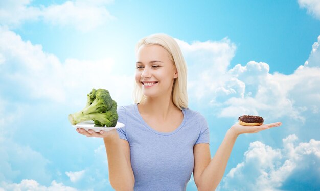Zdrowe Odżywianie, Niezdrowe Jedzenie, Dieta I Wybór Ludzi Koncepcja - Uśmiechnięta Kobieta Wybierająca Między Brokułami I Pączkiem Na Tle Błękitnego Nieba I Chmur