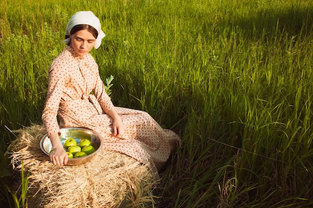 Zdrowe życie na wsi. Kobieta w szaliku siedząca na stogu siana z jabłkami na zielonej łące