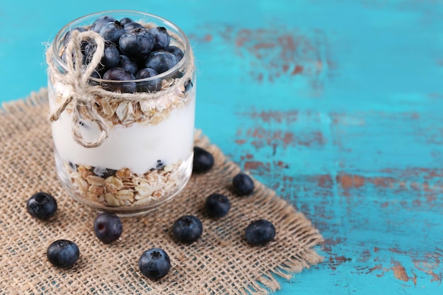 Zdrowy jogurt śniadaniowy z jagodami i musli podawany w szklanym słoju na kolorowym drewnianym tle