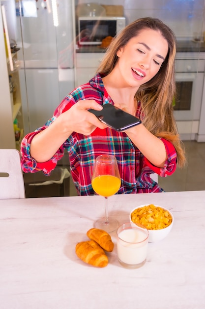 Zdrowy styl życia, kaukaska blondynka pije sok pomarańczowy na śniadanie i robi zdjęcie na portalach społecznościowych