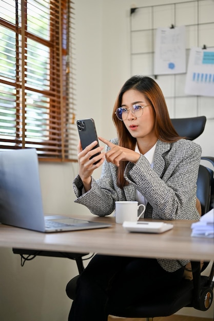 Zdumiona azjatycka bizneswoman używająca smartfona do czytania internetowych plotek