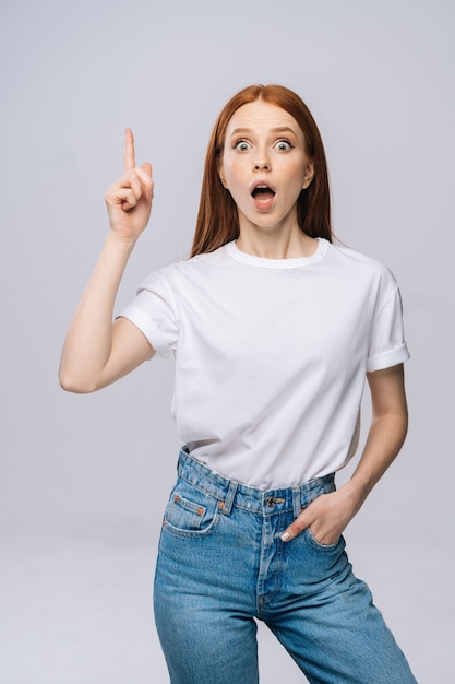 Zdumiona młoda kobieta w koszulce i dżinsowych spodniach, wskazując palcami w górę z otwartymi ustami