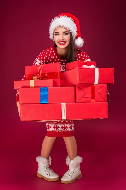 Zdumiona piękna kobieta trzyma wiele prezentów na czerwonym tle. Młoda kobieta w santa hat patrzy na kamerę
