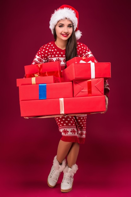 Zdumiona piękna kobieta trzyma wiele prezentów na czerwonym tle. Młoda kobieta w santa hat patrzy na kamerę