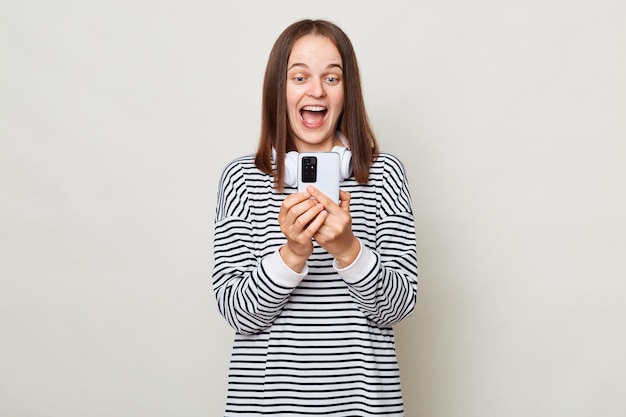 Zdumiona roześmiana brązowowłosa kobieta ubrana w pasiastą koszulę pozuje odizolowana na szarym tle za pomocą telefonu komórkowego czytająca pozytywne wiadomości patrząc na wyświetlacz gadżetu