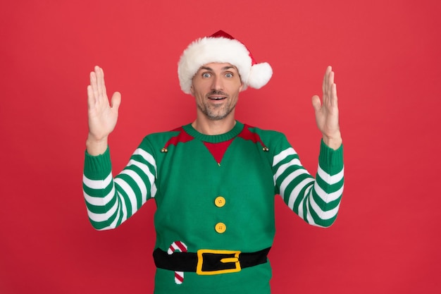 Zdumiony mężczyzna w kostiumie elfa świąteczny facet w kapeluszu świętego mikołaja na czerwonym tle szczęśliwego nowego roku