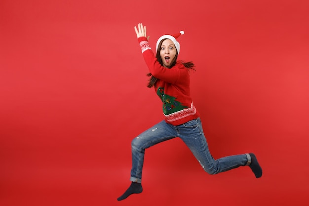 Zdumiony Santa Dziewczyna W Boże Narodzenie Kapelusz Skoki, Wznoszące Się Ręce Rozkładające Nogi, Trzymając Szeroko Otwarte Usta Na Białym Tle Na Czerwonym Tle. Szczęśliwego Nowego Roku 2019 Celebracja Party Wakacje Koncepcja. Makieta Miejsca Na Kopię.
