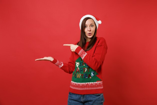 Zdumiony Santa dziewczyna w sweter z dzianiny, Boże Narodzenie kapelusz patrząc zaskoczony, wskazując ręką, palcem na bok na białym tle na czerwonym tle. Szczęśliwego nowego roku 2019 celebracja party wakacje koncepcja. Makieta miejsca na kopię.