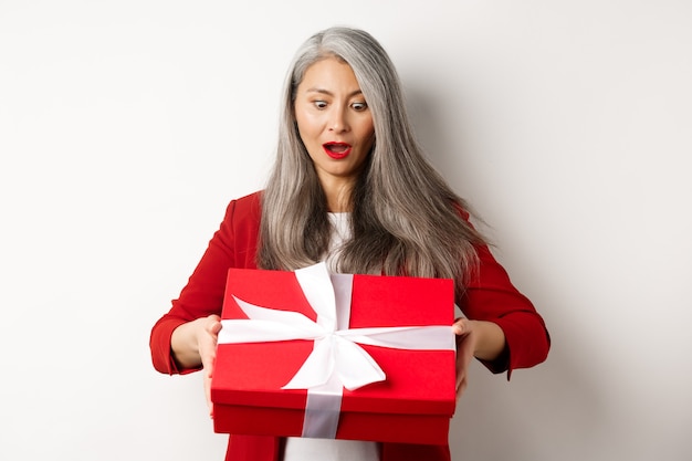 Zdziwiona azjatycka starsza kobieta odbiera prezent na dzień matki, trzymając czerwone pudełko z prezentem i patrząc zdziwiony, białe tło.