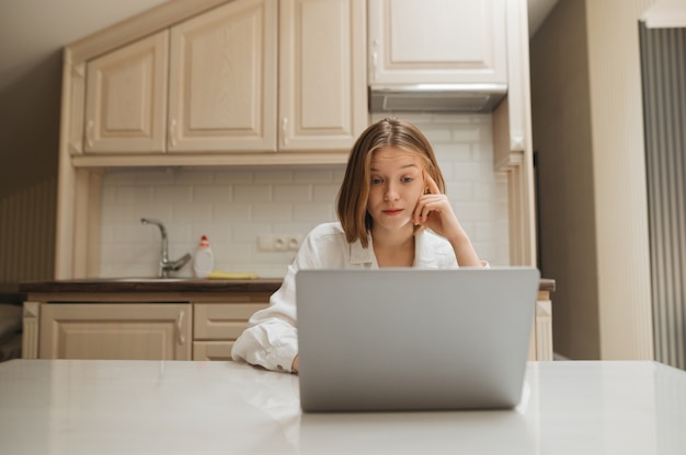 Zdziwiona dziewczyna pracuje na laptopie w domu w kuchni w mieszkaniu
