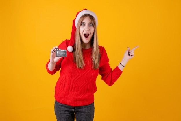 Zdziwiona kobieta trzyma szablon kredytową kartę i wskazuje przy kopii przestrzenią jest ubranym Santa kapelusz