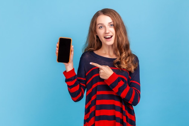 Zdziwiona kobieta ubrana w pasiasty sweter w stylu casual, wskazując na telefon komórkowy z pustym czarnym ekranem na reklamę, która ma zdziwioną ekspresję Kryty studio strzał na białym tle na niebieskim tle