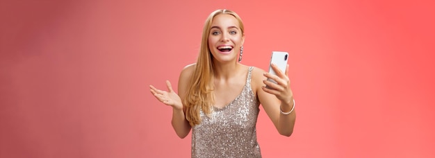 Zdziwiona szczęśliwa urocza blondynka w srebrnej błyszczącej stylowej sukience trzymająca smartfon zdumiona