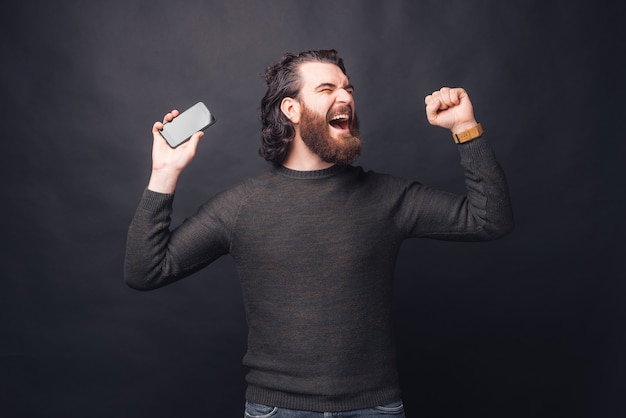 Zdziwiony brodaty mężczyzna w swobodnym stylu świętuje sukces i trzyma smartfon