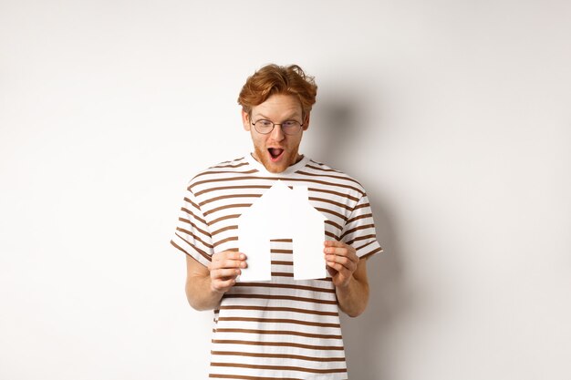 Zdziwiony młody człowiek z rudymi włosami, patrząc z podekscytowaniem na wycinankę z papieru, stojący na białym tle.