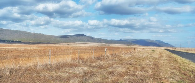 Zebrane grunty rolne obok autostrady w pochmurny dzień Puste pole pszenicy na tle błękitnego nieba Wiejskie rolnictwo z suchymi pastwiskami w pobliżu gór Trawa rosnąca obok pustej drogi w kraju