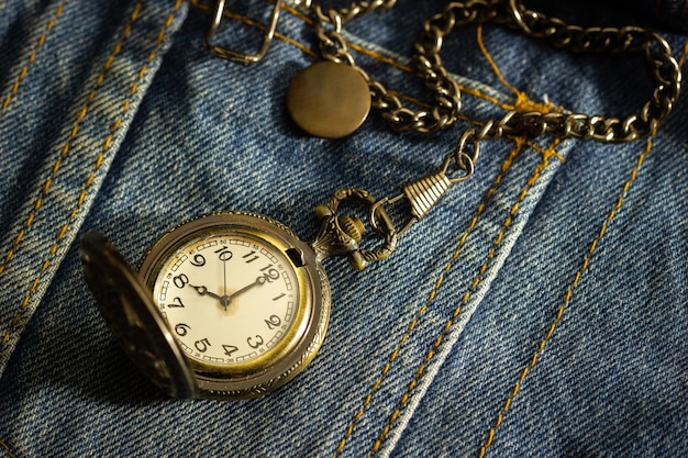 Zegarek kieszonkowy vintage jest umieszczony na starej niebieskiej koszuli dżinsowej, a poranne słońce świeci w prawym górnym rogu. Pojęcie ważności czasu. Zbliżenie i skopiuj miejsce na tekst lub artykuł.