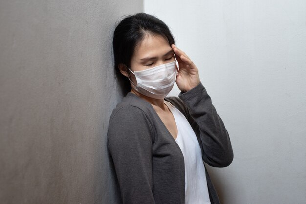 Żeńska Azjatycka młoda kobieta jest ubranym maskę chirurgiczną czuje chorego ból głowy i kaszle opiera na ścianie odizolowywającej na białym tle. Wohan coronavirus zapobieganie wybuchowi. Pojęcie opieki zdrowotnej