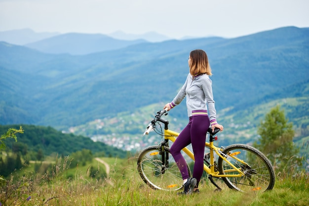 Żeńska cyklista pozycja z żółtym bicyklem na trawie w wierzchołku góra na chmurnym wieczór. Góry, lasy i małe miasto na niewyraźne tło