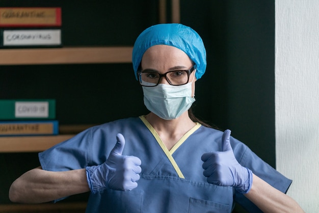Żeński student medycyny w błękitnym ochronnym mundurze i rozporządzalnych rękawiczkach pokazuje aprobata gesta pozycję w biurze