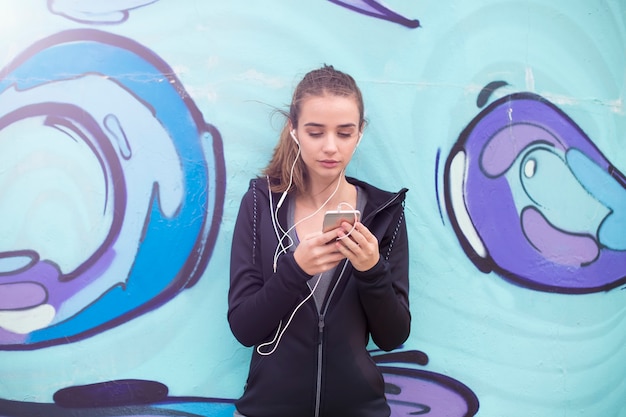 Żeńskiego jogger słuchająca muzyka na telefonie komórkowym przed graffiti ścianą