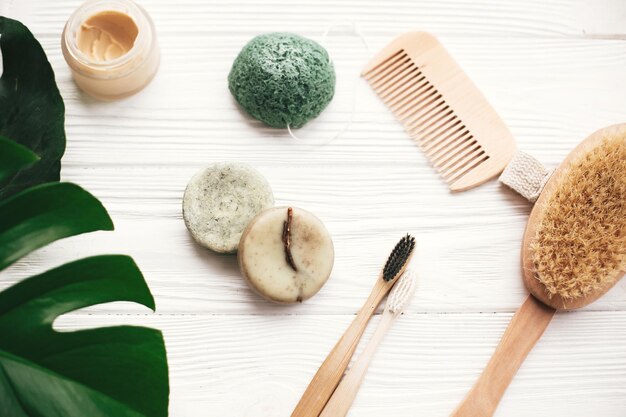 Zdjęcie zero waste flat lay natural solid szampon w kostce bambusowe szczoteczki do zębów drewniana szczotka dezodorant krem i gąbka konjaku na białym drewnie z zielonymi liśćmi monstera produkty ekologiczne bez plastiku