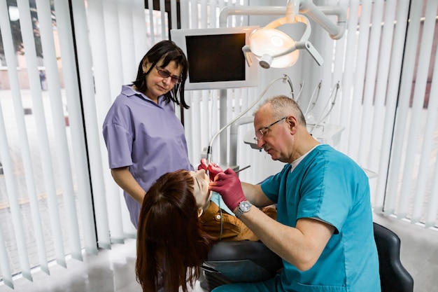 Zespół dentysty medycznego w nowoczesnym gabinecie stomatologicznym robi leczenie zęba dla kobiet młody pacjent. Starszy męski dentysta i jego asystent pracuje w nowożytnej klinice stomatologicznej