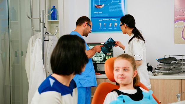 Zdjęcie zespół lekarzy stomatologów analizujący prześwietlenie zębów małej pacjentki z bólem zęba siedzącej na fotelu stomatologicznym rozmawiającej z mamą. lekarz rozmawia z pielęgniarką na temat zdrowia jamy ustnej dziecka.