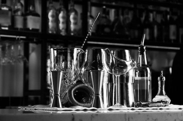 Zestaw akcesoriów barowych do robienia koktajli, znajdujący się na barze, za nim znajduje się regał z likierami i mocnym alkoholem. Różne środki przekazu
