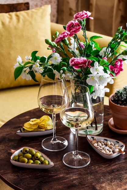 Zestaw aperitifów Dwa kieliszki białego wina i różnorodne przekąski oliwki pistacje chipsy ziemniaczane na drewnianym stole z bukietem kwiatów w przytulnym pokoju