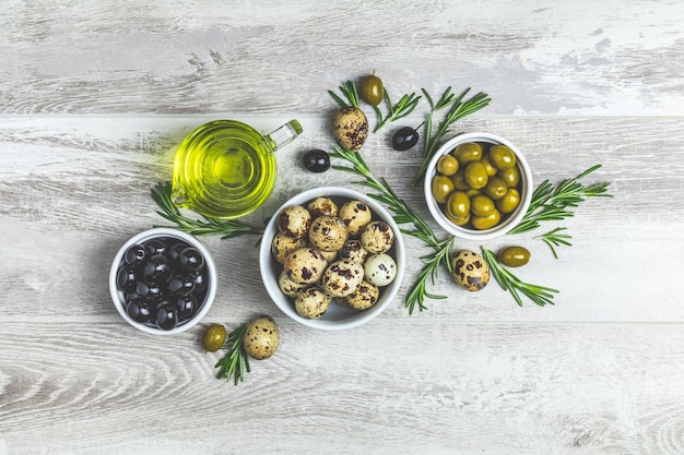 Zestaw jaj przepiórczych z czarnych i zielonych oliwek na talerzach oliwy z oliwek i rozmarynu na jasnoszarym drewnianym tle Widok z góry miejsca