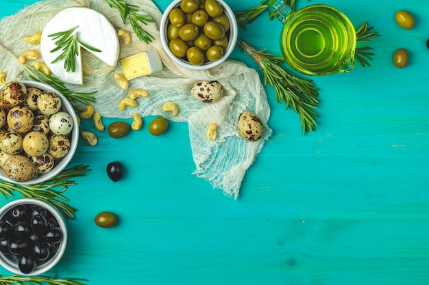 Zestaw serów camembert, czarne i zielone oliwki, jajka przepiórcze na talerzach, oliwa z oliwek i rozmaryn na niebieskim, turkusowym drewnianym stole, widok z góry, miejsce na kopię