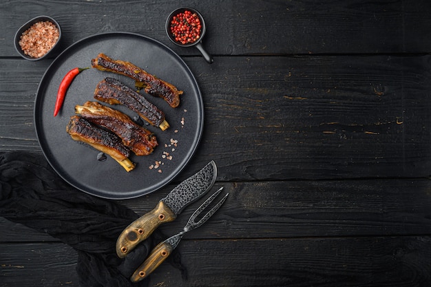 Zdjęcie zestaw żeber wieprzowych z grilla, na talerzu, z nożem do grilla i widelcem do mięsa, na czarnym drewnianym stole, widok z góry na płasko