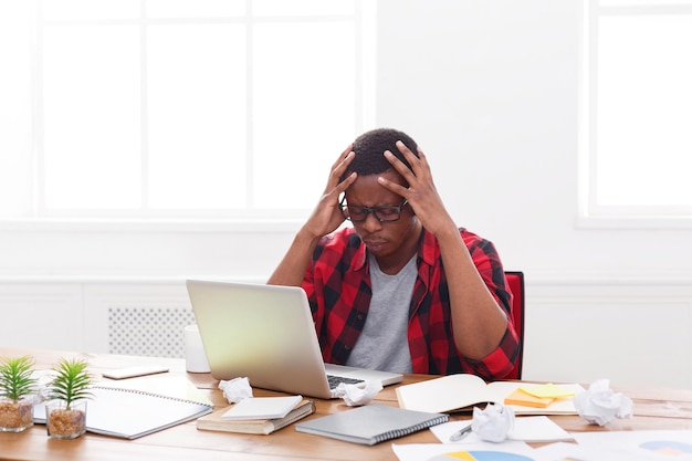 Zestresowany afro-amerykański pracownik z bólem głowy pracujący z laptopem w biurze. Zdenerwowany podczas pracy nad nowym projektem, kopiuj przestrzeń