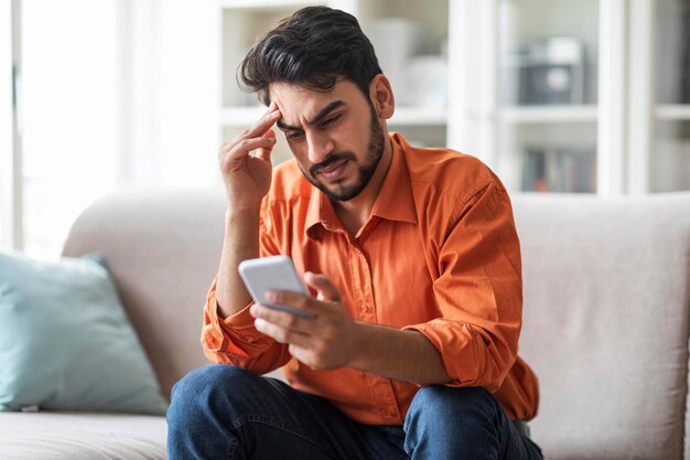 Zestresowany arabski facet siedzi na kanapie w domu trzymając telefon