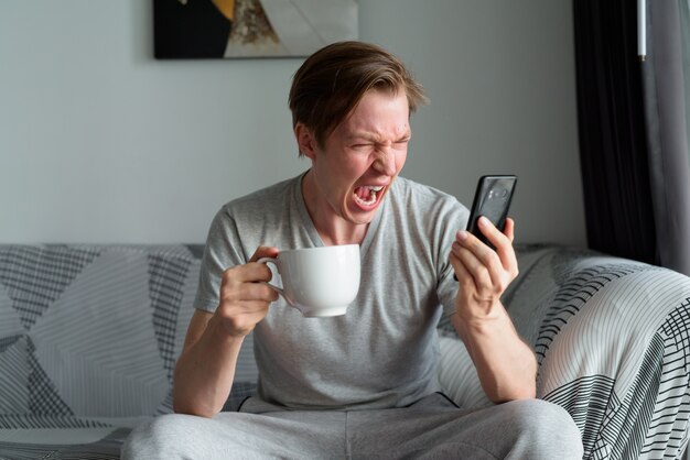 Zestresowany młody człowiek trzyma kawę krzycząc przez telefon w domu