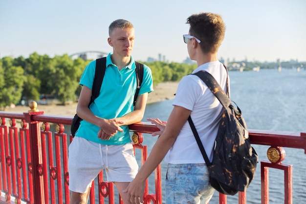 Zdjęcie zewnątrz portret dwóch przyjaciół chłopcy nastolatki 15, 16 lat, rozmawiając, śmiejąc się. faceci stojący na moście nad rzeką w słoneczny letni dzień. młodzież, przyjaźń, komunikacja