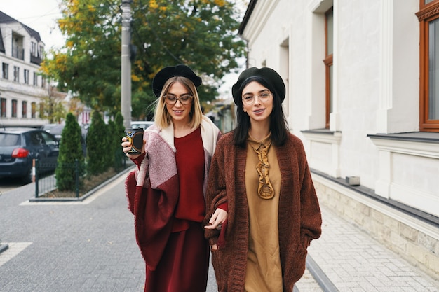 Zewnątrz portret dwóch przyjaciółek. Dziewczyny w zwykłych, ciepłych strojach i okularach spacerujące po mieście w zimnych porach roku i bawiące się na miejskiej ulicy. Miejski styl życia, koncepcja przyjaźni.