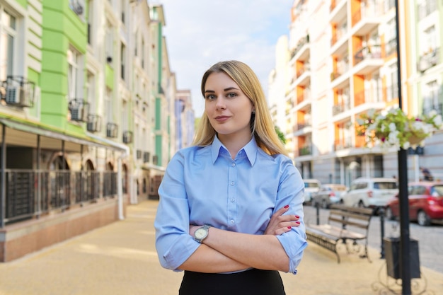 Zewnątrz portret młodej kobiety pewnie biznesu ze skrzyżowanymi rękami, tło ulicy miasta. Kobieta pracownik biurowy w niebieskiej koszuli patrzy na kamerę