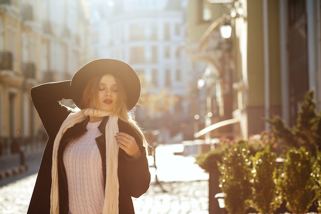 Zdjęcie zewnątrz portret wspaniałej kobiety blondynka na sobie kapelusz, szalik i płaszcz, pozowanie na starej ulicy w blasku słońca. miejsce na tekst