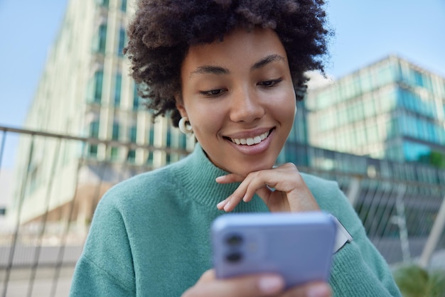 Zewnątrz strzał szczęśliwy kręcone włosy kobieta sprawdza kanał informacyjny na smartfonie uśmiecha się radośnie ubrany w sweter dorywczo trzyma telefon komórkowy pozuje na tle miejskiego. Koncepcja nowoczesnej technologii.