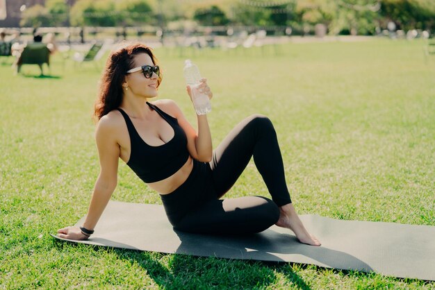 Zewnątrz ujęcie aktywnej szczupłej kobiety pijącej wodę z butelki czuje pragnienie po treningu fitness skoncentrowanym na dystansie nosi przycięty top i legginsy ćwiczy jogę poza koncepcją sportu
