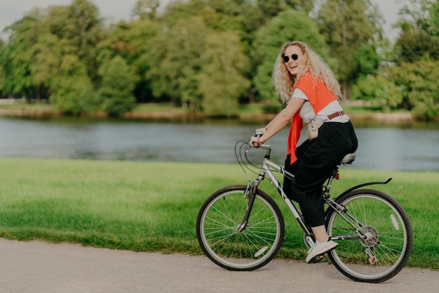 Zdjęcie zewnątrz ujęcie szczęśliwej blondynki kręconej kobiety nosi okulary przeciwsłoneczne, ubrane w swobodne aktywne noszenie, jeździ na rowerze w pobliżu jeziora i zielonych drzew na wsi, spędza wolny czas na zewnątrz lubi swoje ulubione hobby