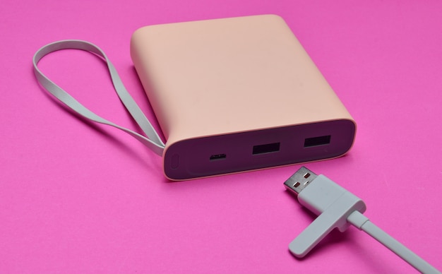 Zewnętrzna bateria do ładowania smartfonów i gadżetów z zbliżeniem kabla USB na różowym tle. Power bank. Nowoczesne technologie.