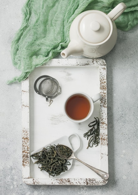 Zielona herbata sypka z zaparzaczem z sitkiem do herbaty i ceramicznym czajniczkiem z filiżanką w drewnianym pudełku z zielonym materiałem na jasnym tle